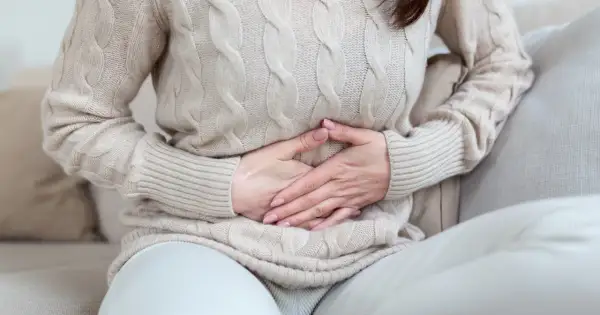 Kobieta w beżowym swetrze trzyma się za brzuch, odczuwając bolesne rozpychanie dziecka w brzuchu.